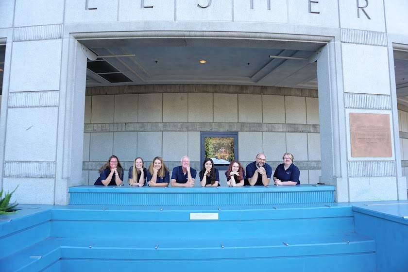 A group of 8 people smile and post behind a blue bench and against building exterior